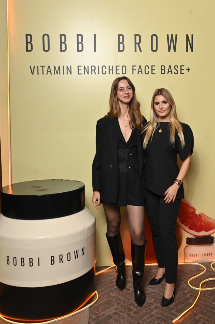 Bobbi Brown etkinliğinde bir araya gelmiş, şık ve modern giyimli kadın konuklar, ellerindeki içecekleri tutarak sıcak gülümsemelerle birbirleriyle keyifli sohbetler ediyor, samimi ve canlı bir atmosferde sosyalleşiyorlar.