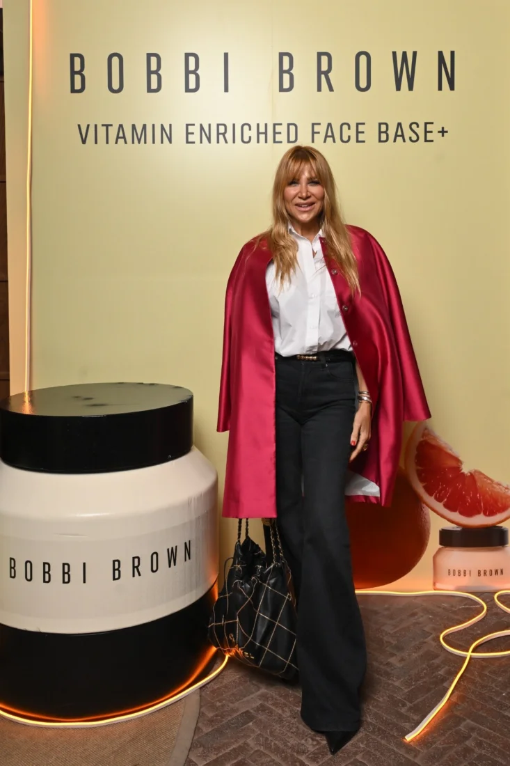Bobbi Brown etkinliğinde bir araya gelen, yüzlerinde samimi gülümsemelerle makyaj ürünlerinin bulunduğu masanın önünde sohbet eden ve keyifli anlar geçiren, modern ve şık giyimli kadın konuklar, sosyal ve enerjik bir atmosferde kameraya bakıyor.