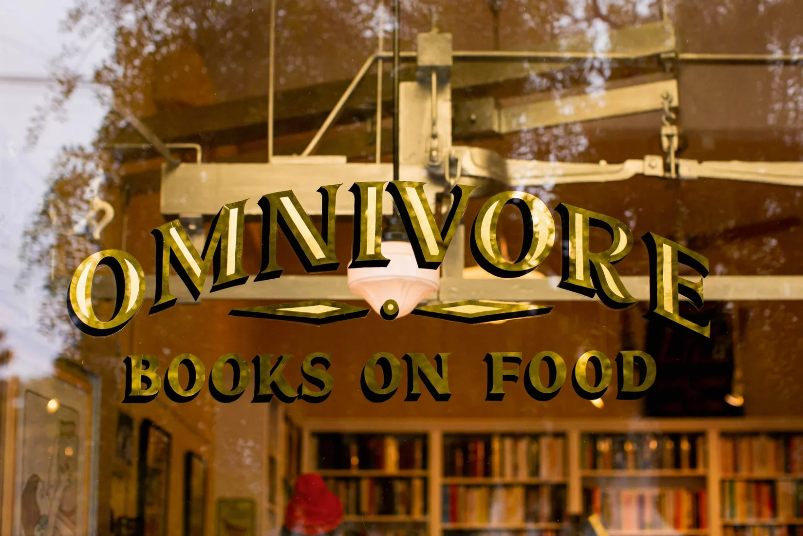 San Francisco'daki Omnivore Books on Food adlı gastronomi kitapçısının ön camında, dışarıdaki yansımalarla birlikte parlayan, altın ve siyah tonlarda yazılmış büyüleyici logosu belirgin bir şekilde duruyor; içeride ise raflara dizili nadir ve koleksiyonluk yemek kitapları, okurları eşsiz lezzet hikâyelerine davet ediyor.