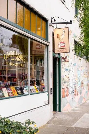San Francisco'daki Omnivore Books on Food'un altın renkli harflerle yazılı adını gösteren vitrini, içerideki zengin yemek kitabı koleksiyonunu yansıtırken, asma tabelasındaki kitaplar üzerindeki horoz figürü ve yan duvardaki renkli gastronomi temalı duvar resmi, bu mekanın dünyanın en özel gastronomi kitapçıları arasındaki yerini vurguluyor.