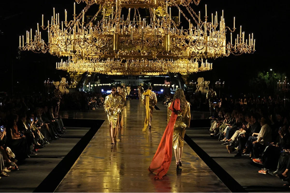 Uzun boylu, kısa saçlı bir model, Paris Moda Haftası kapsamında Saint Laurent'in görkemli defilesinde, dramatik ışıklarla aydınlatılmış bir podyumda modern ve sofistike bir tasarımla kendine güvenli adımlarla yürürken.