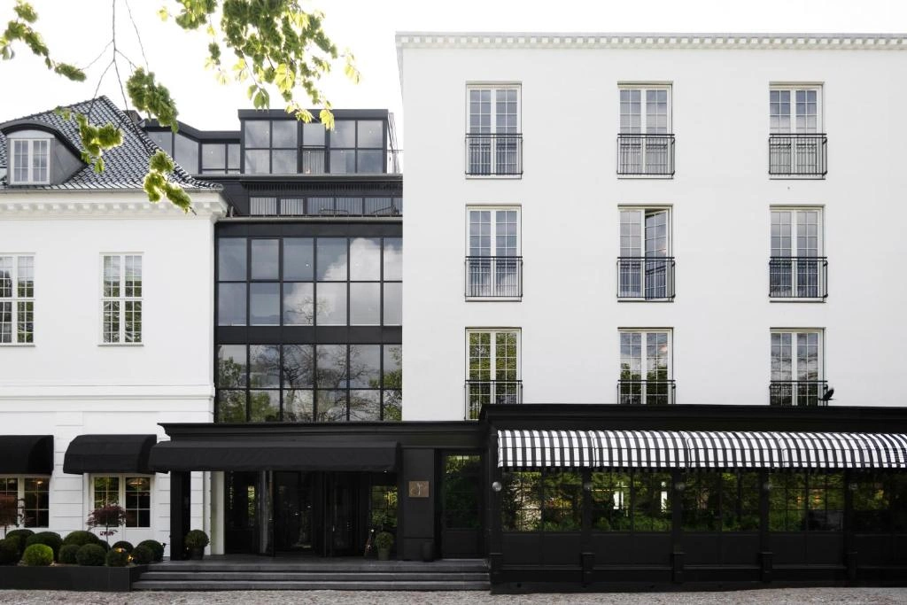 Hellerup'un sakin atmosferindeki Park Lane Copenhagen otelinin, solda kiremit çatılı klasik beyaz binayı, ortada geniş cam cepheli modern yapıyı ve sağda ferforje balkonlu minimalist beyaz bloğu birleştiren dış görünümü, Kopenhag'ın İskandinav şıklığını siyah-beyaz detaylarla gözler önüne seriyor.