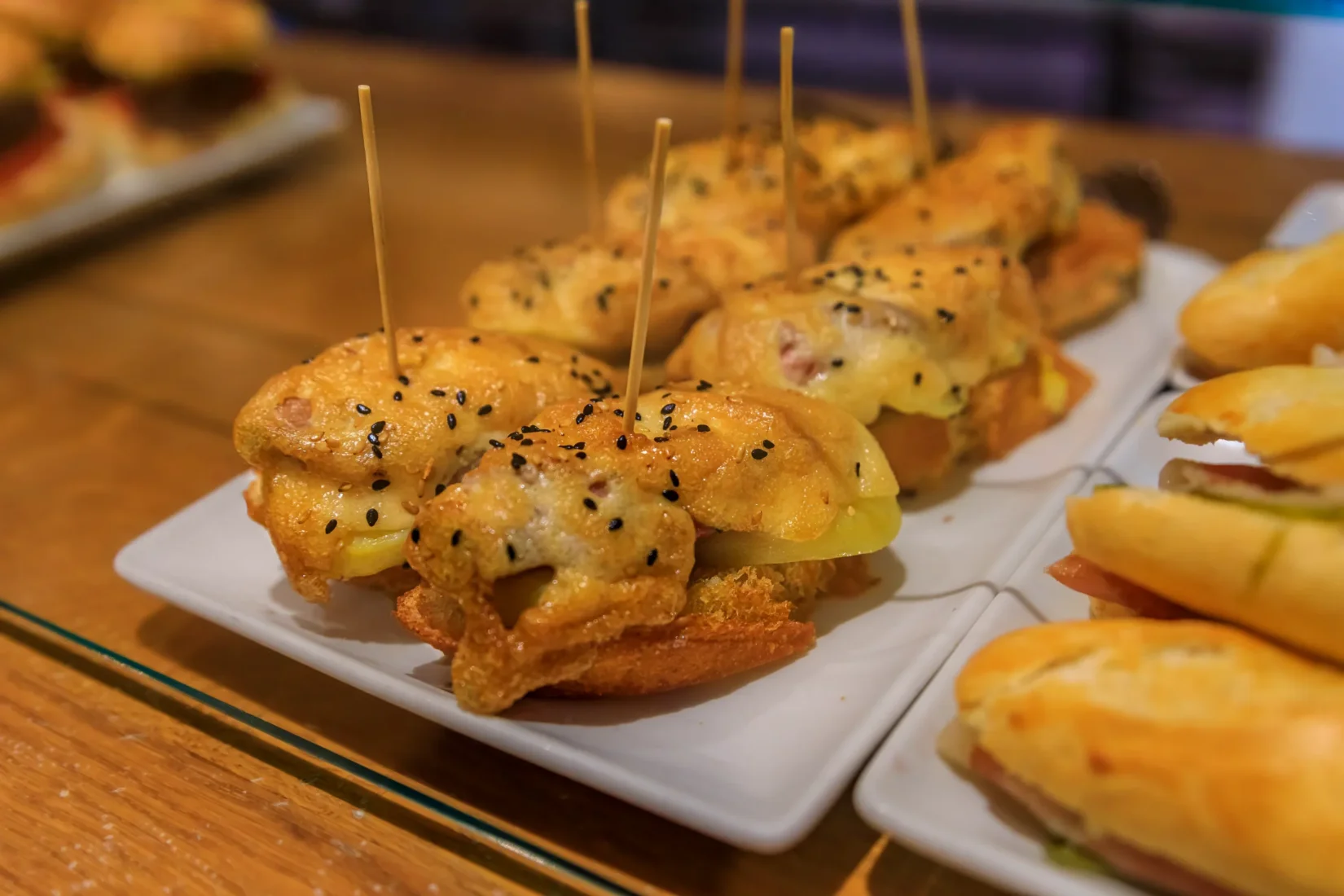 Basque mutfağının geleneksel ve lezzetli atıştırmalıkları olan pintxosları gösteren bu görselde, üzerine farklı deniz ürünleri, peynirler ve sebzeler eklenerek hazırlanmış, kürdanlarla sabitlenmiş, rengarenk ve iştah açıcı birçok küçük ekmek diliminin özenle dizildiği zengin bir sunum masası dikkat çekiyor.