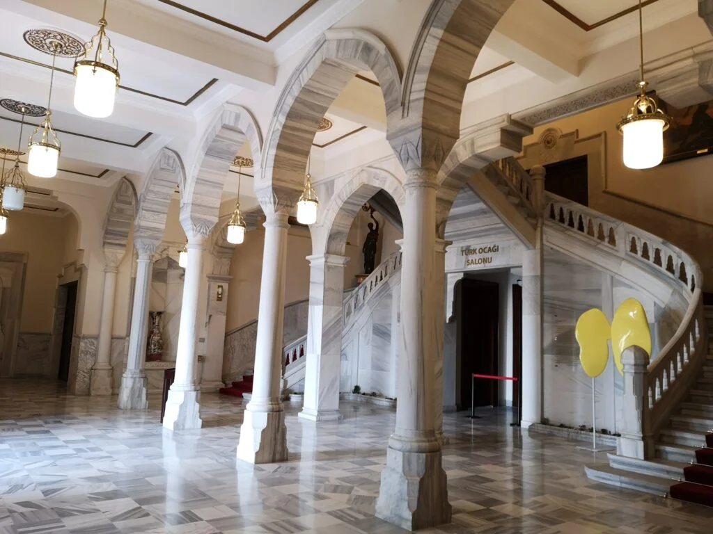 Ankara Resim ve Heykel Müzesi'nin yüksek tavanlı, geniş bir salonunda, duvarları çeşitli tablolarla süslenmiş, orta alanda bir heykelin görüldüğü mimari detaylarla dolu, aydınlık bir iç mekan.