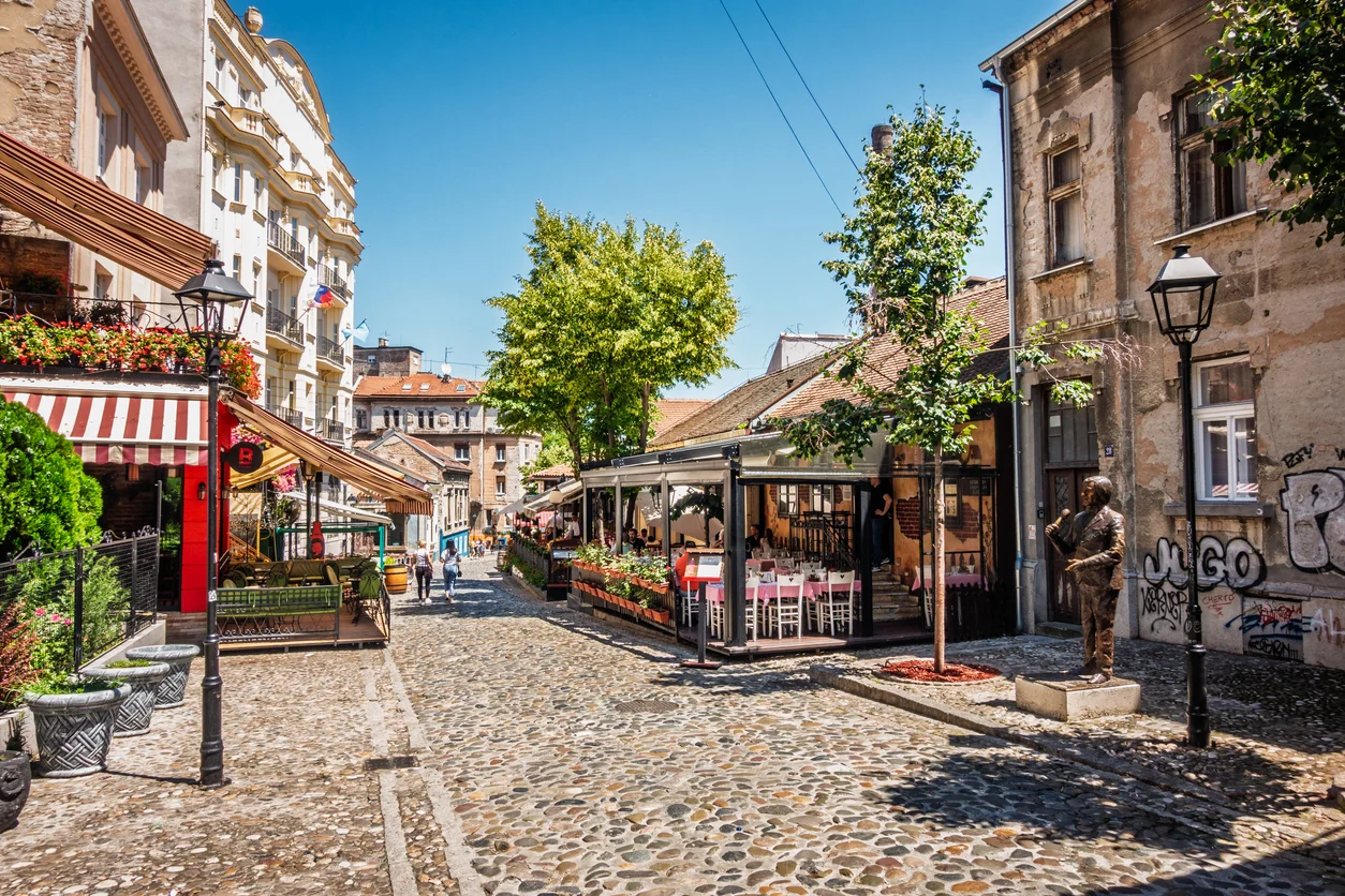 Belgrad'ın tarihi ve Bohem ruhunu yansıtan Skadarlija bölgesinde, ağaçların hafifçe sararmaya başladığı Arnavut kaldırımlı bir sokakta, canlı ve hareketli bir atmosferde açık hava masalarında oturan, yürüyen ve sohbet eden insanlarla dolu, çiçeklerle süslü geleneksel binaların sıralandığı pitoresk bir cadde görünümü.