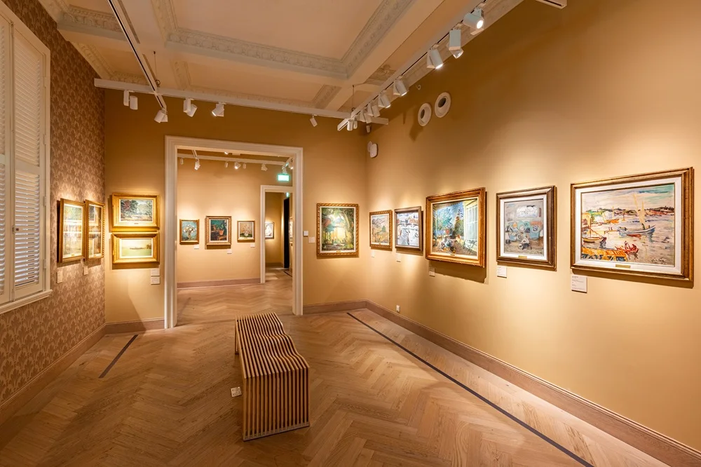 Sergide, ziyaretçiler duvardaki çeşitli ressamlar tarafından yapılmış tabloları dikkatle inceliyor, sanat eserlerinin detaylarına odaklanırken modern bir sanat galerisinin aydınlık ve ferah ortamı dikkat çekiyor.