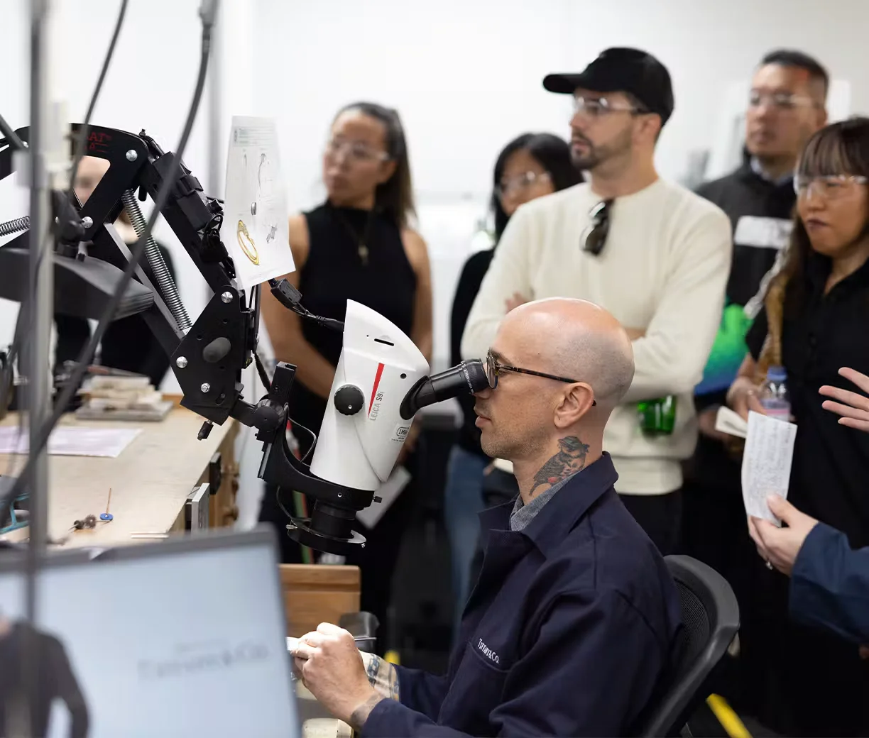 Bir Tiffany & Co. atölyesinde gözlüklü ve dövmeli bir usta, Leica mikroskop altında titizlikle bir mücevher parçasını işlerken, LVMH’nin Les Journées Particulières etkinliği kapsamında lüksün sırlarını keşfetmeye gelen bir grup ziyaretçi merakla onu izliyor, bu an ışıltılı tasarımların ardındaki değerli zanaatkarlığı ve el emeğini gözler önüne seriyor.
