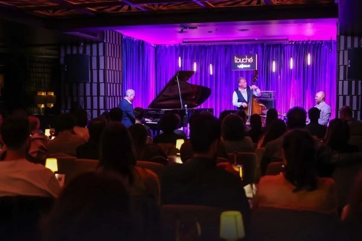 İstanbul'un caz kulüpleri arasında popüler bir yer olan Touché'de, mor ışıklandırılmış sahnede piyano ve kontrbas eşliğinde canlı bir caz performansı sunan sanatçıları, loş bir ortamda oturan geniş bir dinleyici topluluğu ilgiyle izliyor.