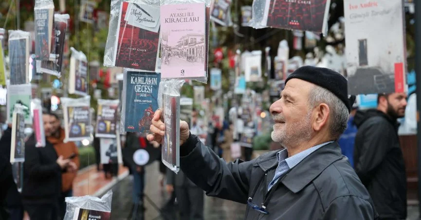 Gözlükleri ve içten gülümsemesiyle Uğur Batı, arkasında kitap dolu rafların hafifçe bulanık göründüğü bir mekanda, yeni kitabı olması muhtemel, dikkat çekici kapaklı bir eseri iki eliyle tutarak okurlarına adeta merhaba diyor ve bu görsel, yazarın edebi kariyerine dair bir içeriği pekiştiriyor.