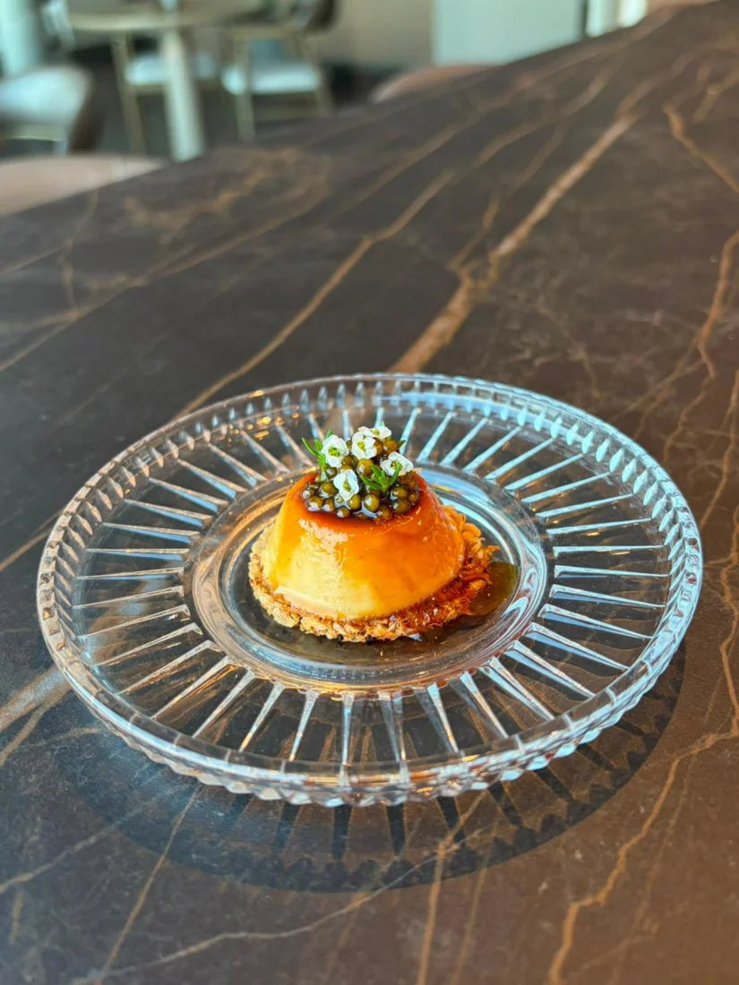 Beyaz bir ramekin içinde servis edilen, parlak sarı renkli pürüzsüz kremalı dokuya sahip ve etrafından süzülen parlak karamel sosuyla zenginleştirilmiş iştah açıcı bir creme caramel tatlısı, üzerinde zarif bir nane yaprağı ile sunuluyor.