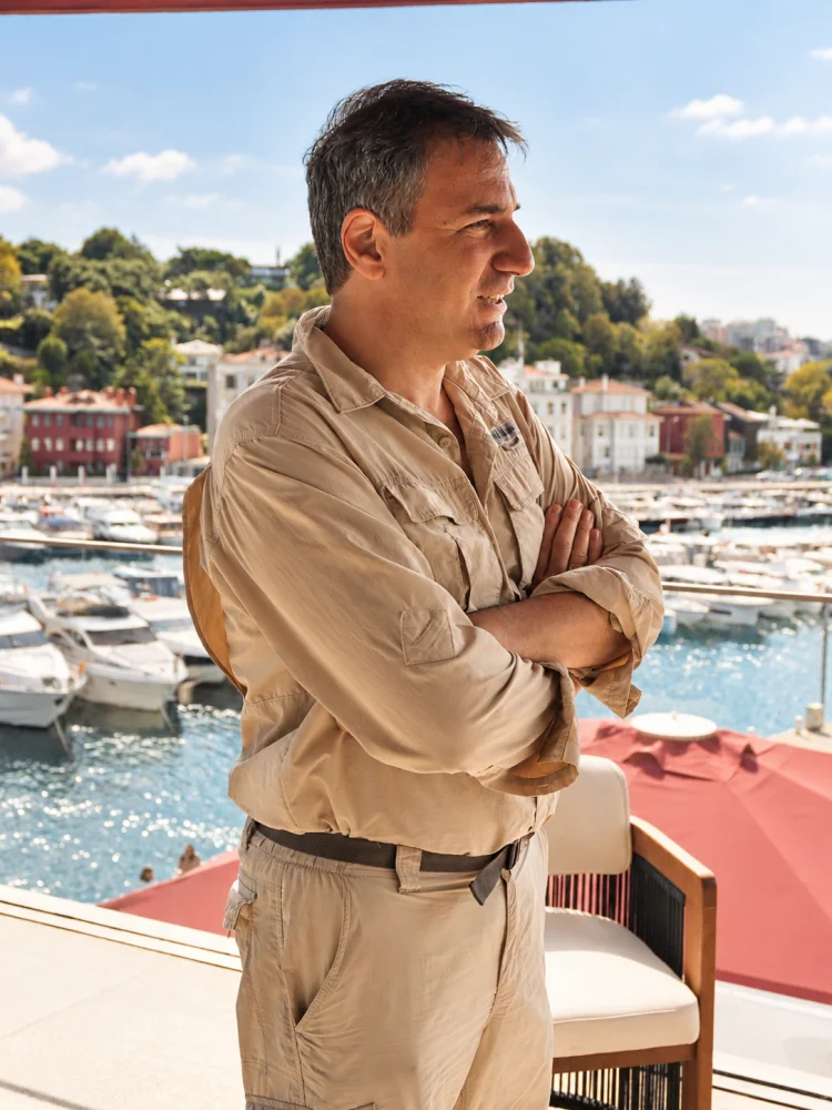 Yat tasarımcısı Mehmet Aziz Göksel, atölyesinde büyük bir yat modeline veya tasarımına odaklanmış bir şekilde dururken, çevresindeki çeşitli tasarım araçları ve prototiplerle dolu çalışma ortamı, onun titiz ve yaratıcı sürecini gözler önüne seriyor.